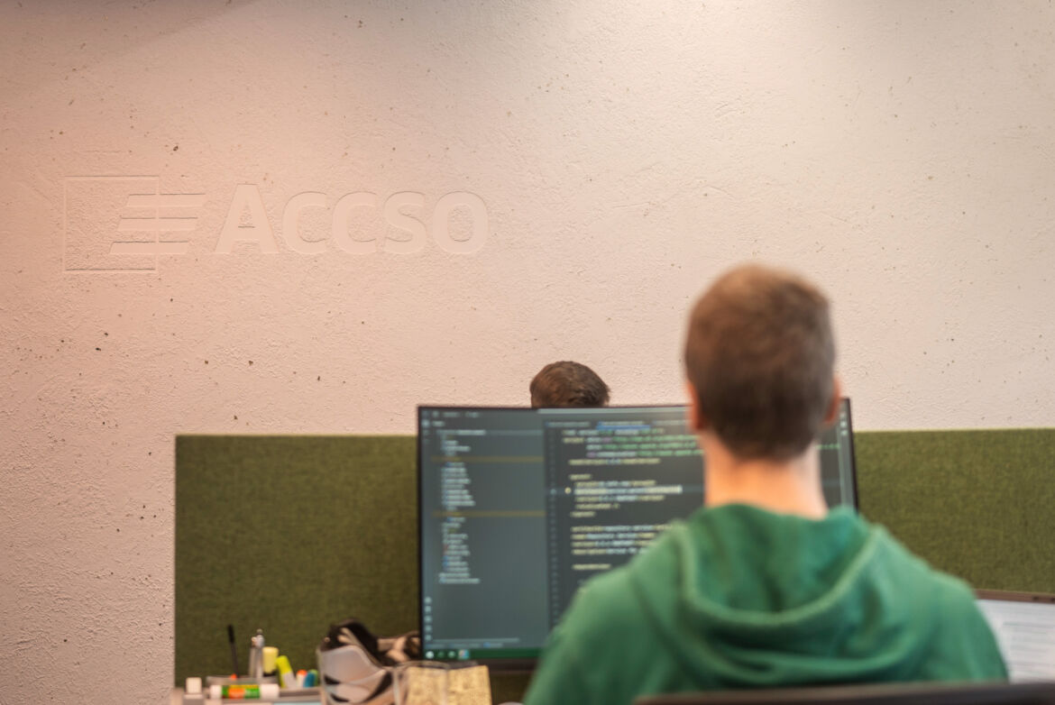 Ein Mann im grünen Pullover sitzt mit dem Rücken zum Betrachter gewandt vor einem Bildschirm, auf dem Code zu sehen ist.